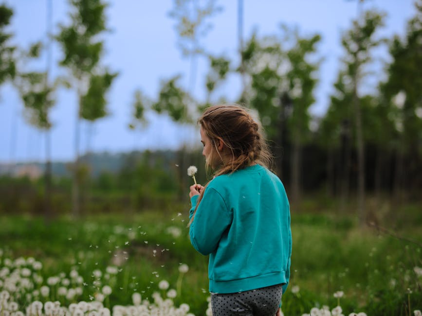 A young girl in a meadow blowing a dandelion under a clear sky in Italy, capturing childhood innocence.