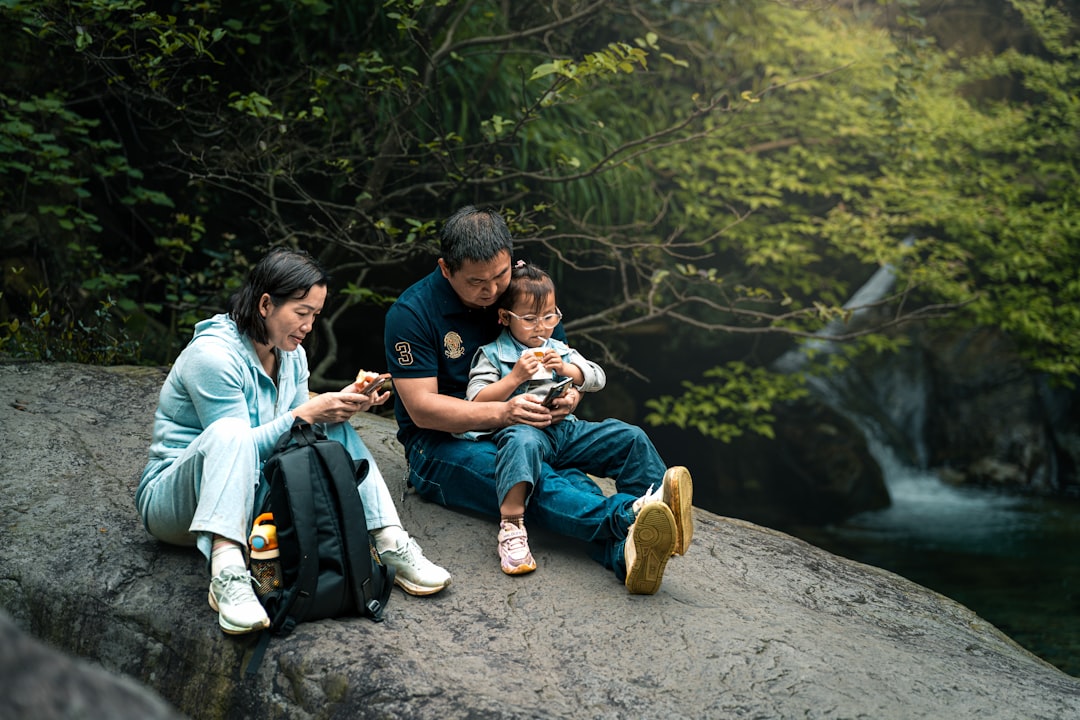 Family relaxing on a rock, using phones by a waterfall.